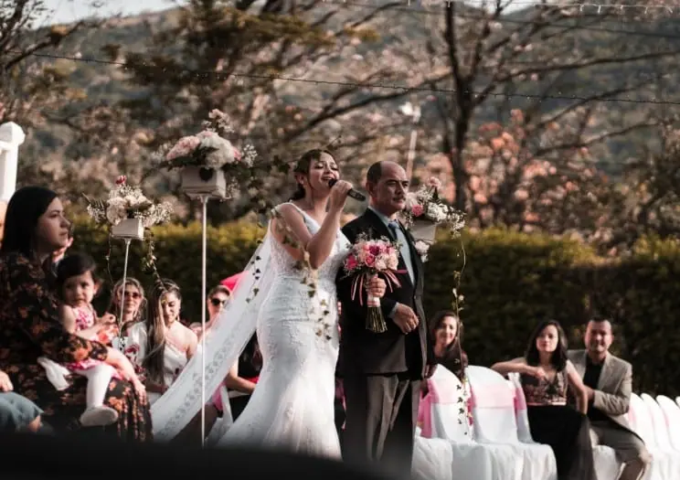 Bride in a veil and groom with guests watching an outdoor ceremony under cherry blossom trees