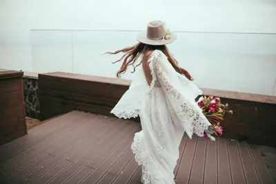 Woman in a boho lace dress and hat walking on a wooden deck holding a bouquet